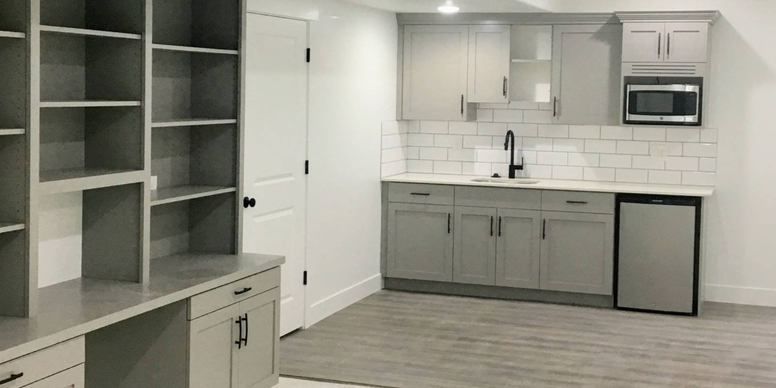 basement finishing building codes saratoga springs, utah: what homeowners need to know 1 A modern white and grey basement kitchenette featuring a grey cabinet in the foreground, situated next to a door, showcasing the results of obtaining basement finishing permits in Saratoga Springs, Utah.